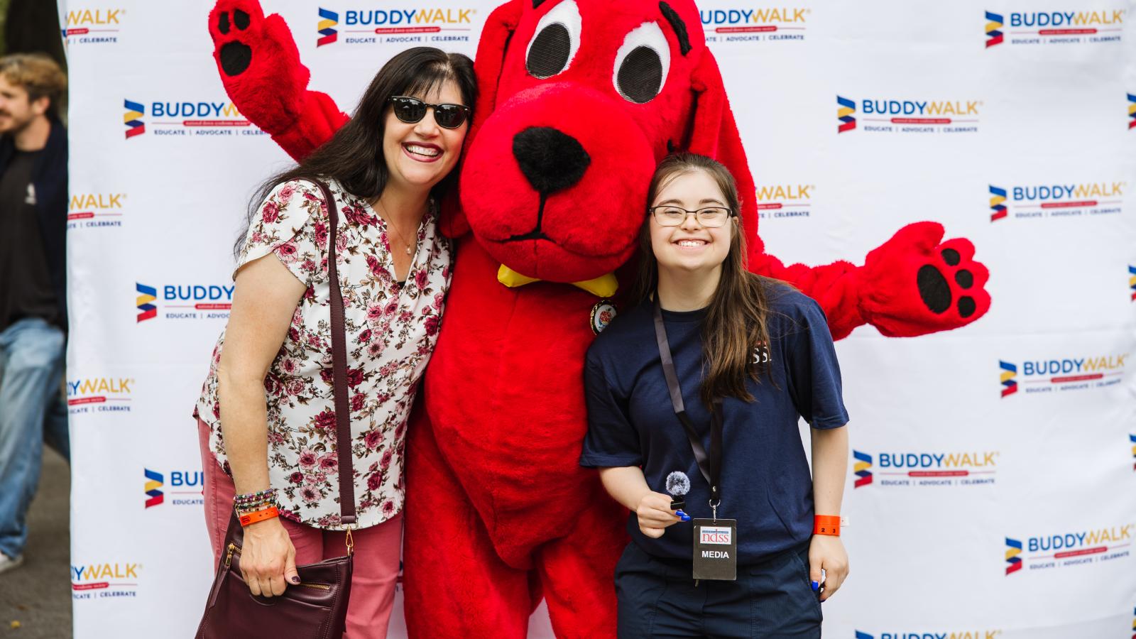 A woman in sunglasses and a floral top stands with her arm around Clifford, who has one paw raised. Beside them, a girl with Down syndrome, wearing glasses and a navy T-shirt with an NDSS logo and a lanyard with a "Media" badge, smiles at the camera. All three stand in front of a white backdrop with the repeating Buddy Walk logo.