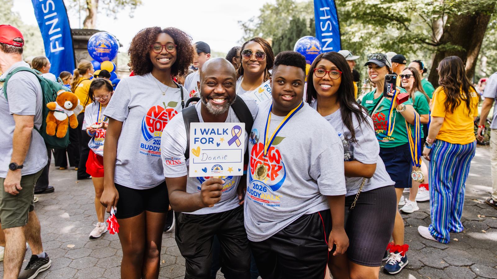 A group of five people stand together outdoors near blue "Finish" flags and blue balloons. A young woman with curly hair and glasses, a man with a beard holding a sign that reads "I walk for Donnie," a woman in sunglasses, a young man with Down syndrome wearing a gold medal, and another woman all wear matching gray T-shirts printed with a red apple design and "New York City 2026 Buddy Walk, Educate | Advocate | Celebrate."