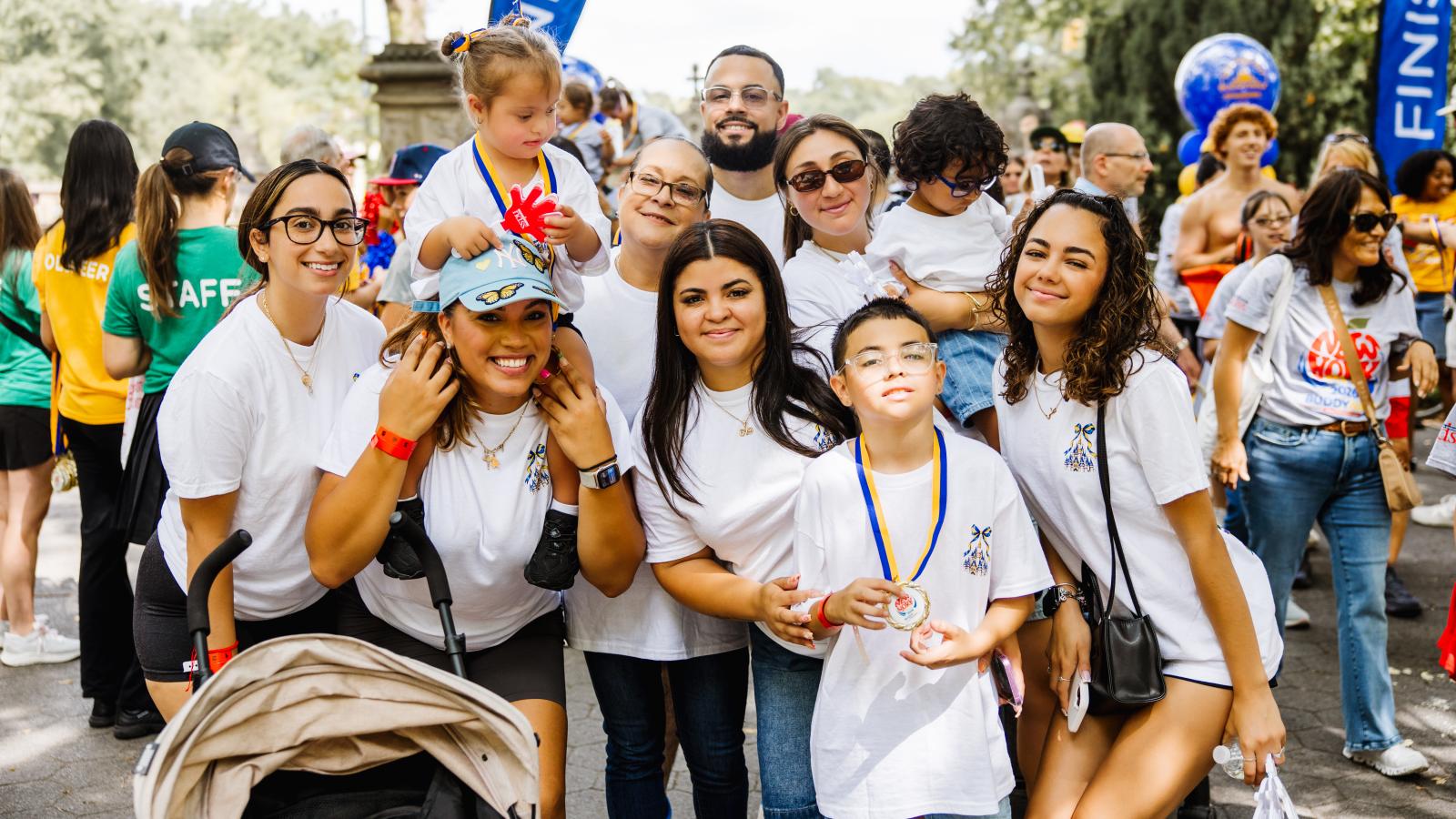 A large group poses outdoors near a blue "Finish" flag banner. A girl with Down syndrome sits on a woman's shoulders; a girl stands nearby. Several adults and children, including a boy with Down syndrome wearing glasses and a gold medal, all wear matching white T-shirts with a small castle logo on the chest. A tan stroller sits in the foreground.