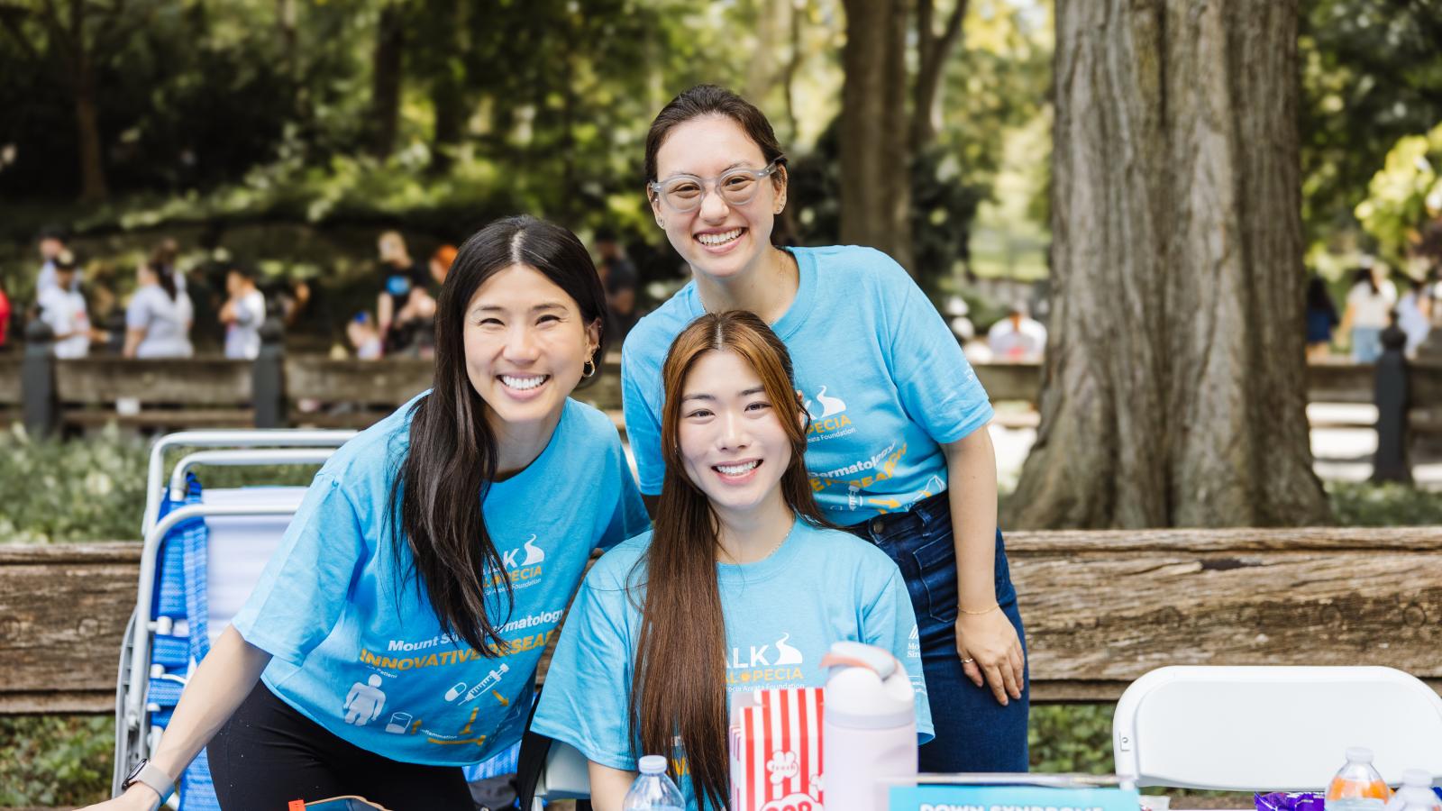 Three women stand behind a table at an outdoor event, all wearing matching light blue T-shirts printed with "Alk alopecia" and "Mount Sinai Dermatology innovative research" alongside small icons of a syringe, pill bottle, and figure. On the table in front of them are red-and-white striped popcorn boxes, water bottles, and a light blue sign reading "Down syndrome dermatology clinical trial interest form."