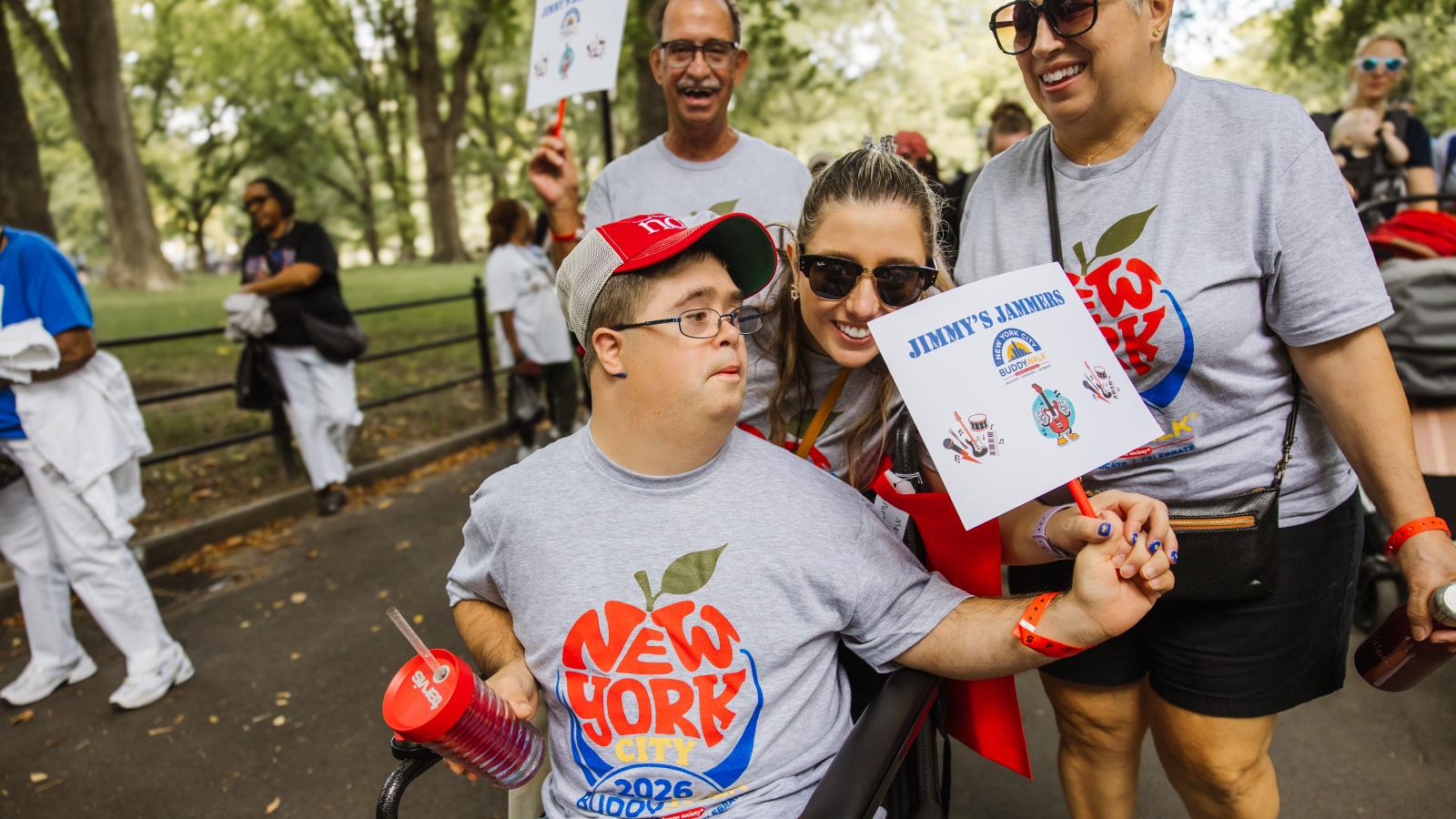 A man with Down syndrome wearing glasses, a red cap, and a gray T-shirt printed with "New York City 2026 Buddy Walk" sits in a mobility chair holding a red water bottle. A woman in sunglasses leans down beside him holding a sign that reads "Jimmy's Jammers" with a New York City Buddy Walk logo and small illustrated music-note and instrument icons. Another woman and man, both in matching gray T-shirts, stand behind them.