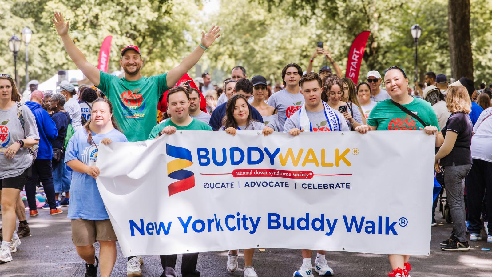 A group of people walk together outdoors holding a white banner that reads "Buddy Walk, National Down Syndrome Society, Educate | Advocate | Celebrate, New York City Buddy Walk." A man in a green T-shirt and red cap raises both arms in celebration behind the banner. Several people with Down syndrome are among the group, wearing gray T-shirts printed with a red apple design and "New York."