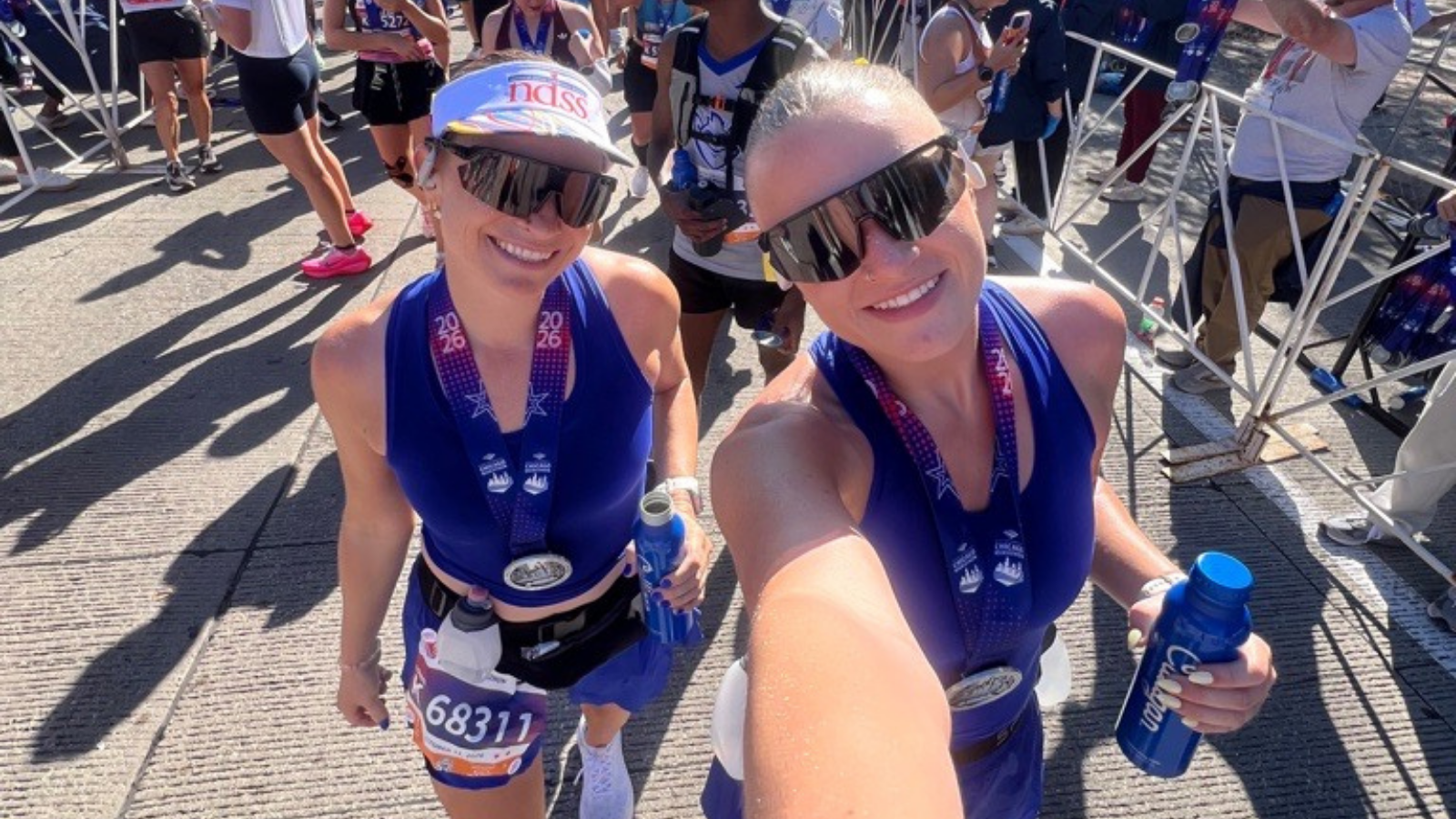 Two smiling women wear royal blue sleeveless tops, blue skirts, black sunglasses, and 2026 Chicago Marathon finisher medals on purple ribbons. The woman on the left wears a white NDSS visor and a race bib reading 68311, and holds a blue water bottle. The woman on the right takes the selfie and holds a blue bottle. Runners and metal barricades are behind them.