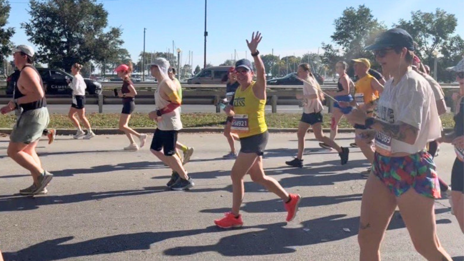 A woman wearing a yellow NDSS tank top, gray shorts, a navy cap, red-tinted sunglasses, and bright orange-red sneakers waves with one hand while running. Her bib reads 68921. She is surrounded by other runners on a sunny road, with a guardrail and sailboat masts in the distance.