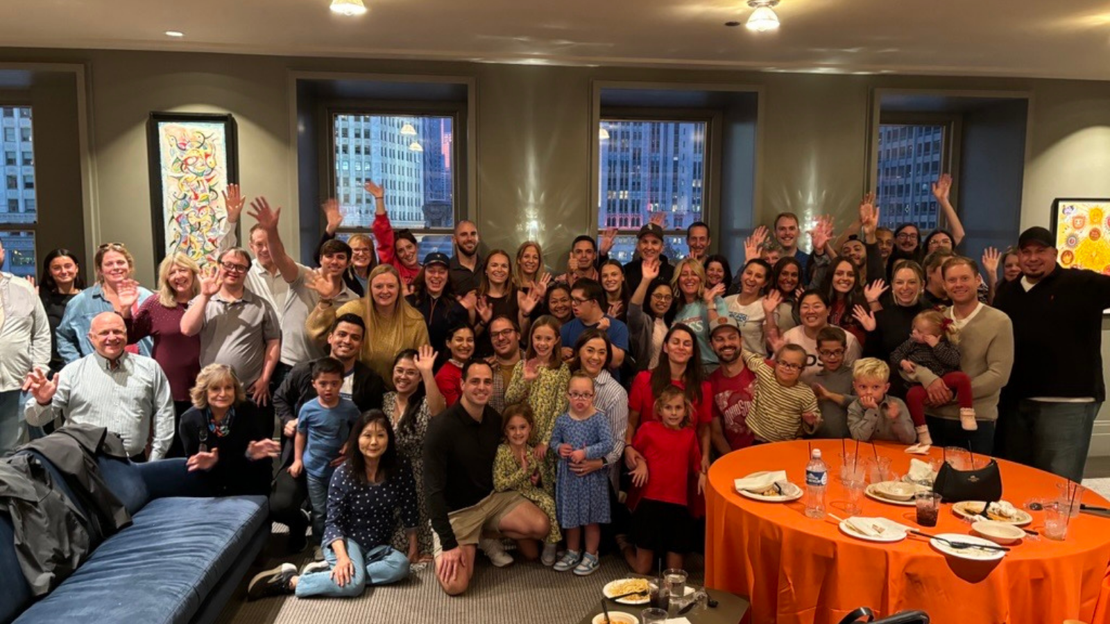 More than 50 adults and children pose and wave for a group photo in a room with large windows overlooking city buildings. A few individuals in the photo have Down syndrome. An orange tablecloth covers a round table with plates and cups on the right, and a blue couch is on the left. Colorful abstract paintings hang on the walls.