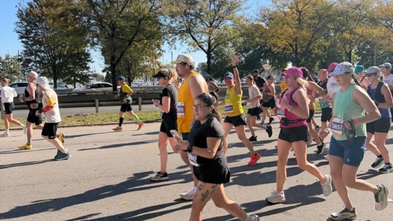 A woman in a yellow NDSS tank top, black shorts, and bright red sneakers waves with one hand while running among a crowd of marathon runners on a sunny, tree-lined road. A man in a yellow tank top and a woman in a black dress run in front of her. Sailboat masts are visible in the background.