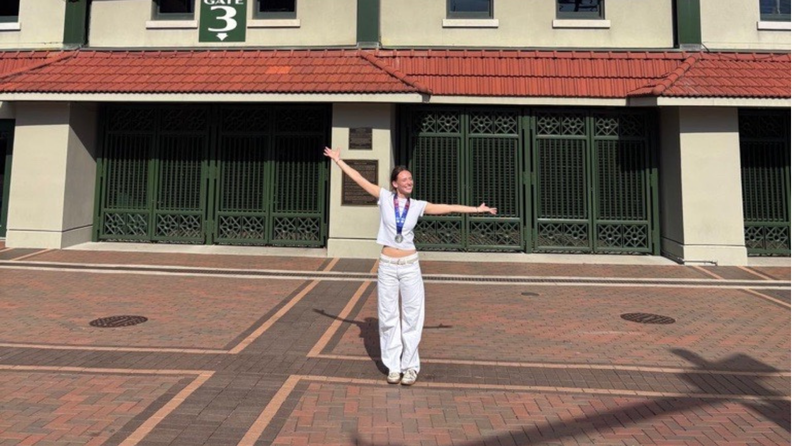 A woman stands with her arms stretched wide in front of a building with a red tile roof and dark green gates. A green sign above reads Gate 3. She wears a white cropped T-shirt, white wide-leg pants, white sneakers, and a finisher medal on a blue ribbon. She stands on a red brick plaza in front of Wrigley Stadium and is smiling.