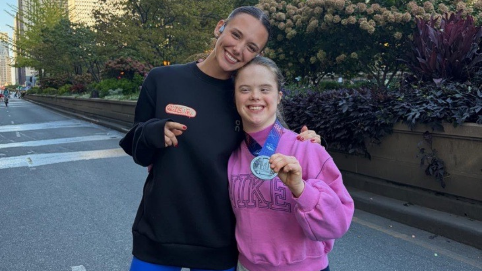 Two women pose close together, smiling, on a closed city street lined with trees and shrubs. The woman on the left wears a black oversized sweatshirt with a small red patch and points toward the camera. The woman on the right has Down syndrome, wears a pink Nike sweatshirt, and holds up a silver finisher medal on a blue ribbon.