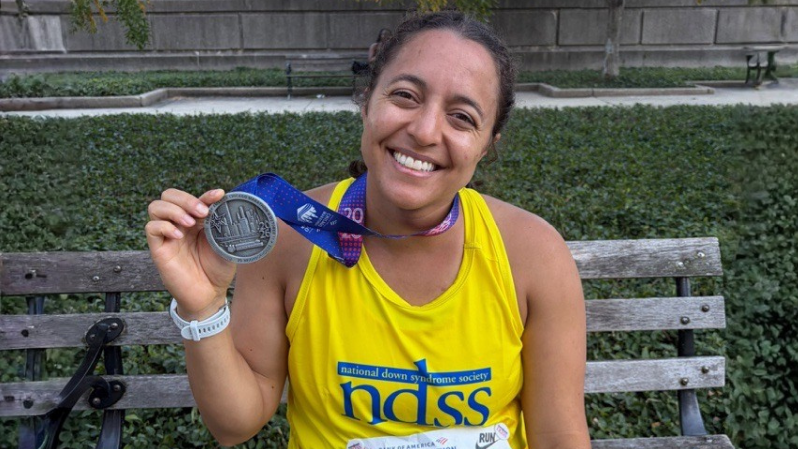 A smiling woman sits on a wooden park bench in front of a green hedge, holding up a silver marathon medal on a blue and purple ribbon. She wears a bright yellow tank top with the words National Down Syndrome Society above large blue NDSS letters, and a white watch.