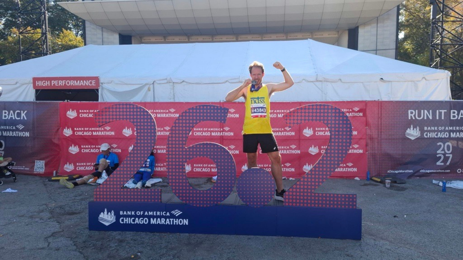 A man in a yellow NDSS tank top, black shorts, and race bib H43843 flexes one arm and holds a medal to his mouth. He stands in front of a large purple and red dotted 26.2 sign on a blue base reading Bank of America Chicago Marathon. Behind him are a red banner wall, a white tent, and a red sign reading High performance.