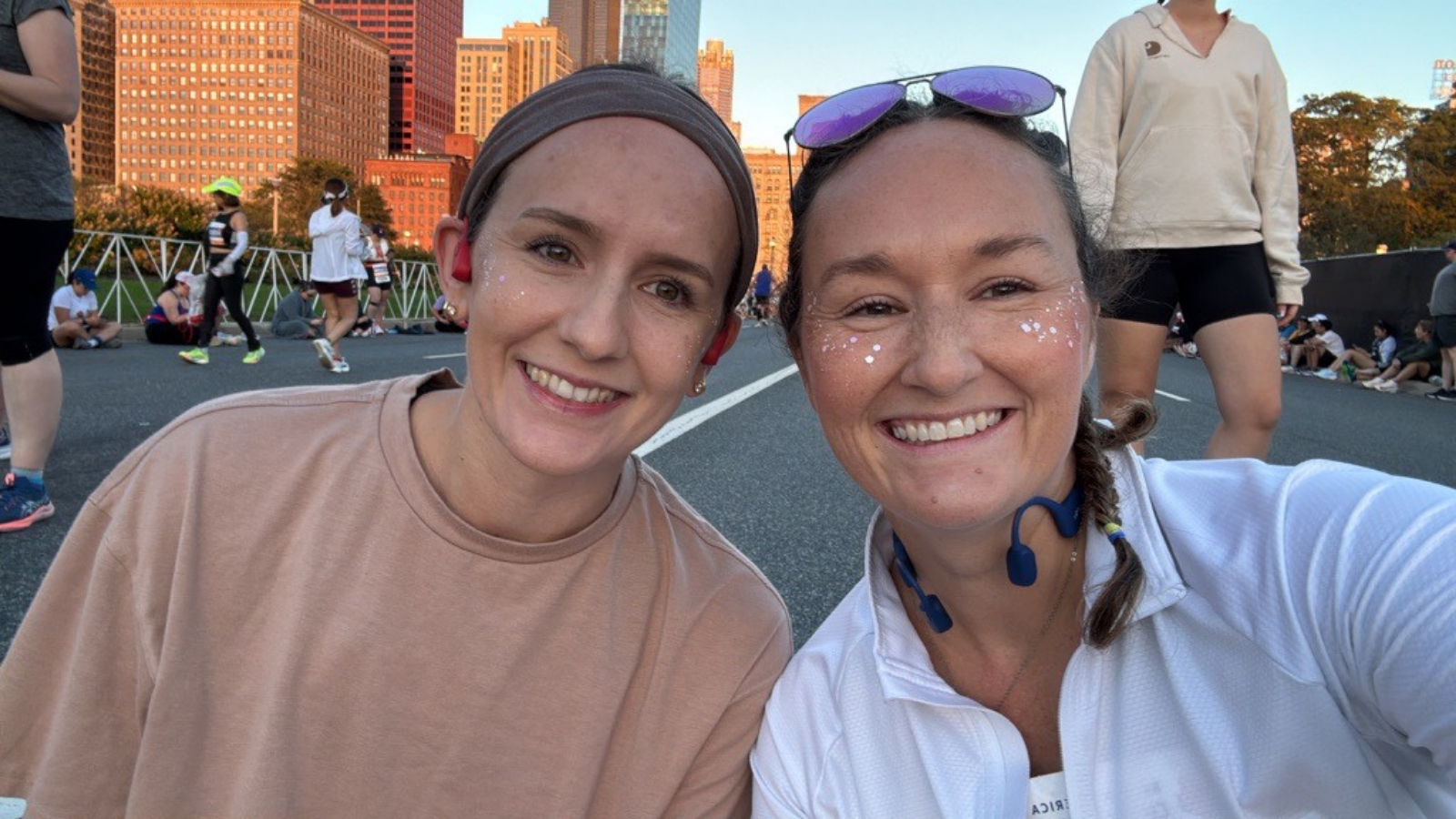 Two smiling women pose for a selfie on a closed city street at sunset, with tall brick buildings behind them. The woman on the left wears a tan T-shirt and a brown headband. The woman on the right wears a white quarter-zip top, purple sunglasses on her head, blue headphones, and a side braid. She has glitter on her cheeks.
