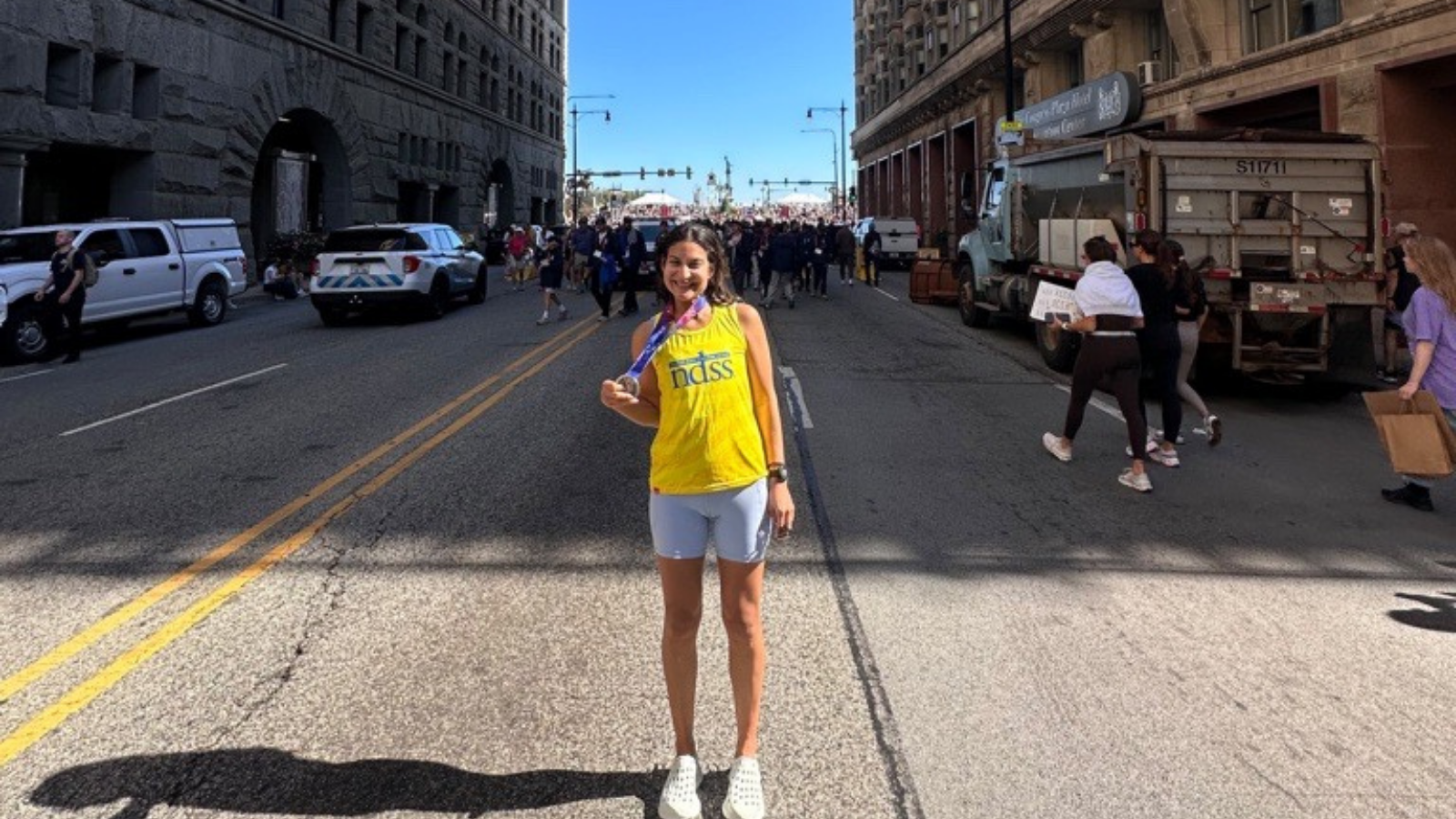 A smiling woman stands in the middle of a street in downtown Chicago holding a finisher medal on a blue and purple ribbon. She wears a yellow NDSS tank top, light blue shorts, and white clogs. Double yellow lines run down the street, with tall stone buildings, a police vehicle, a dump truck, and pedestrians behind her.