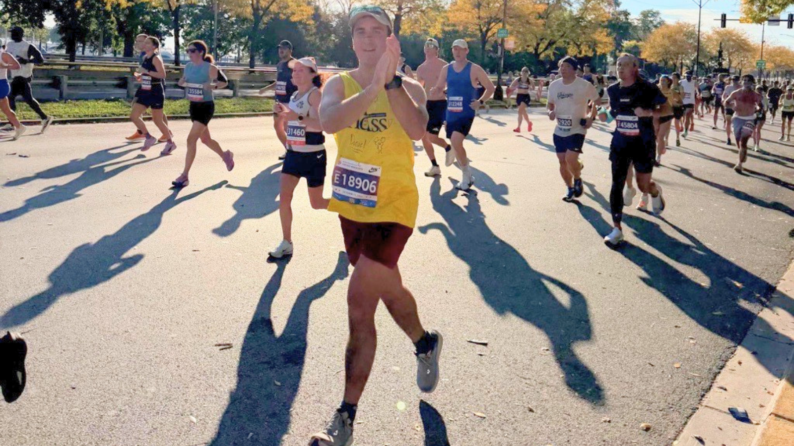 A man in a yellow tank top with NDSS written on it in blue, maroon shorts, a gray cap, and bib E18906 claps while running a race. Other runners in the street are behind him, with trees in yellow and orange fall colors along the course.