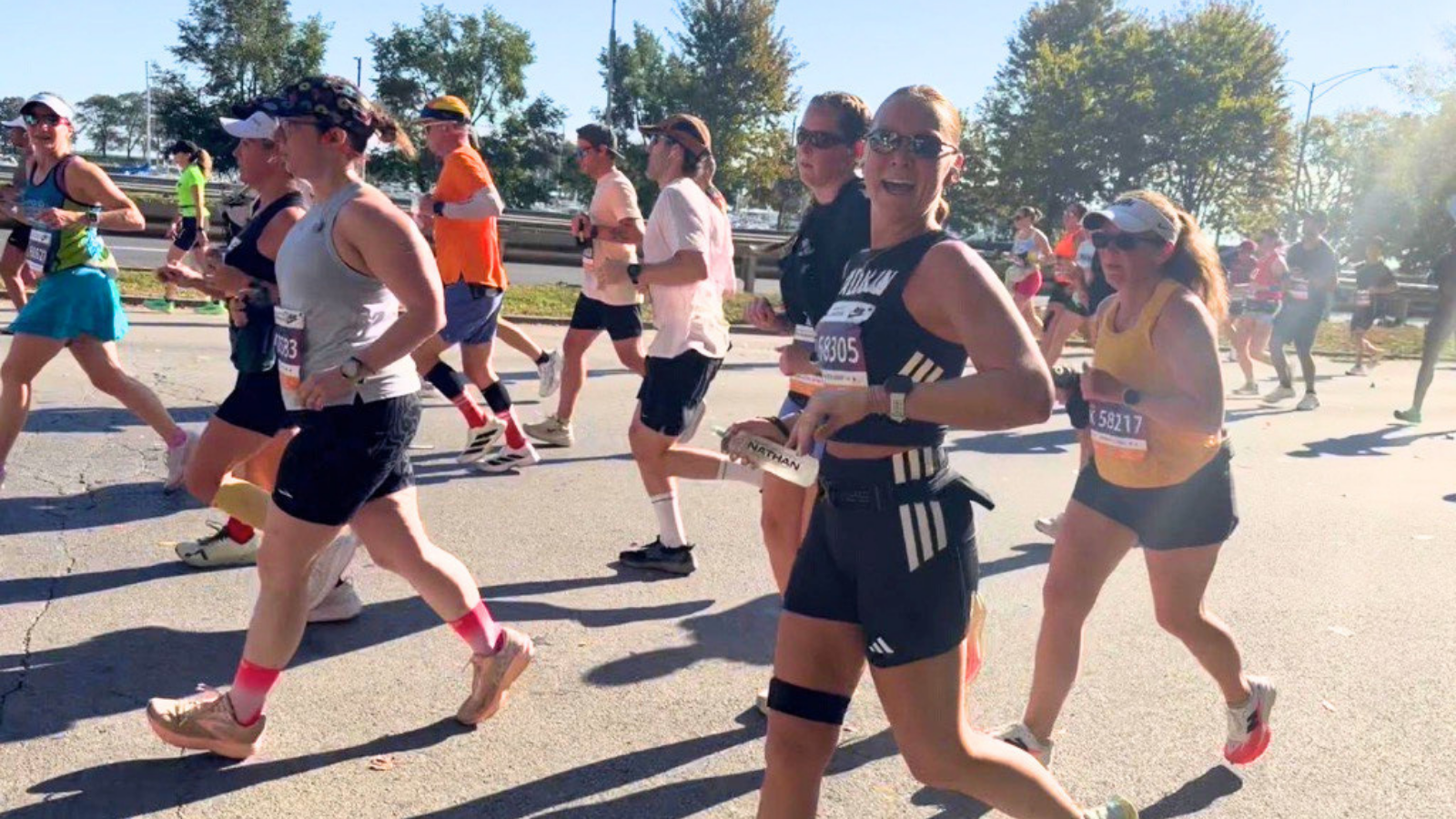 A smiling woman looks toward the camera while running in a crowd of marathon runners. She wears a black sleeveless top with a race bib reading 68305, black Adidas shorts, sunglasses, and a black knee brace, and holds a water bottle. Runners in tank tops and shorts surround her on a sunny, tree-lined road.