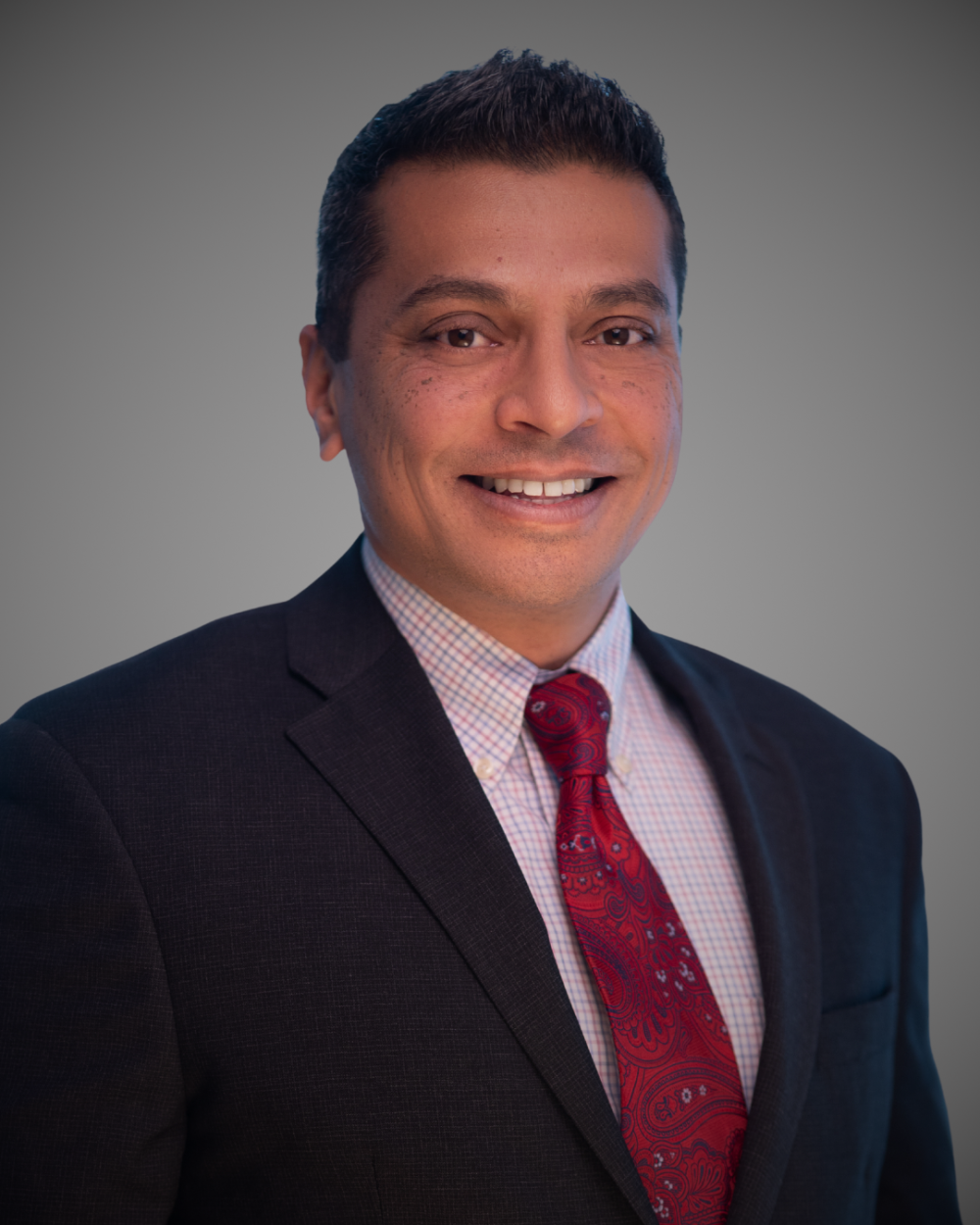 Hasan Shah smiling in his headshot with a blue suit, red button down and red paisley tie