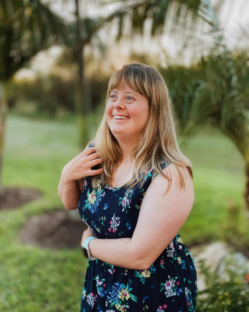 Photo of Candace Whiting, a woman with Down syndrome. She has dirty-blonde hair and wears a black dress with colorful flowers on it. She stands outside with palm trees in the background.