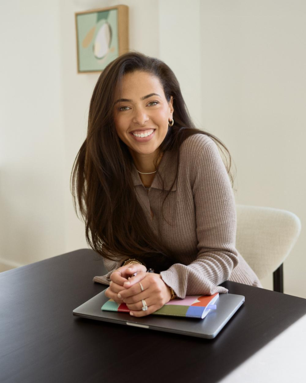 Photo of Ciara Siegel, a woman with dark brown hair, wearing a tan sweater. She wits at a desk with a laptop and notebook.