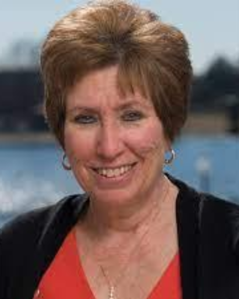 Photo of Dr. Amy Lewanda, a woman with short brown hair, wearing a black top. She stands outside in front of a body of water.