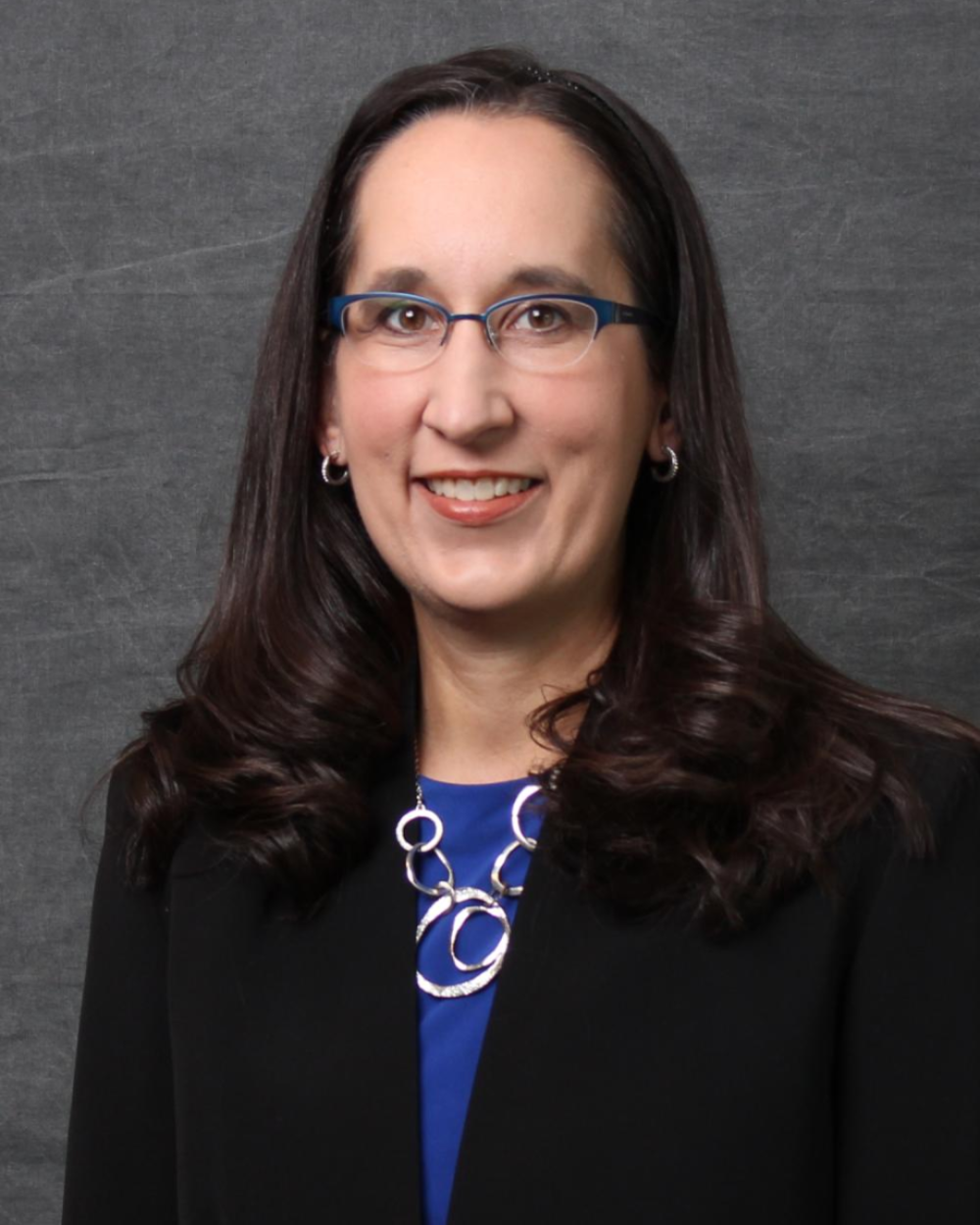 Headshot of Dr. Heather Anderson, a woman with dark brown hair, wearing glasses, a blue blouse, black blazer, and a necklace with gold rings.