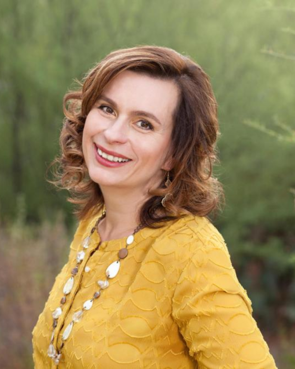Photo of Dr. Jamie Edgin, a woman with brown hair, wearing a peach blouse. She outside in front of tall grasses.