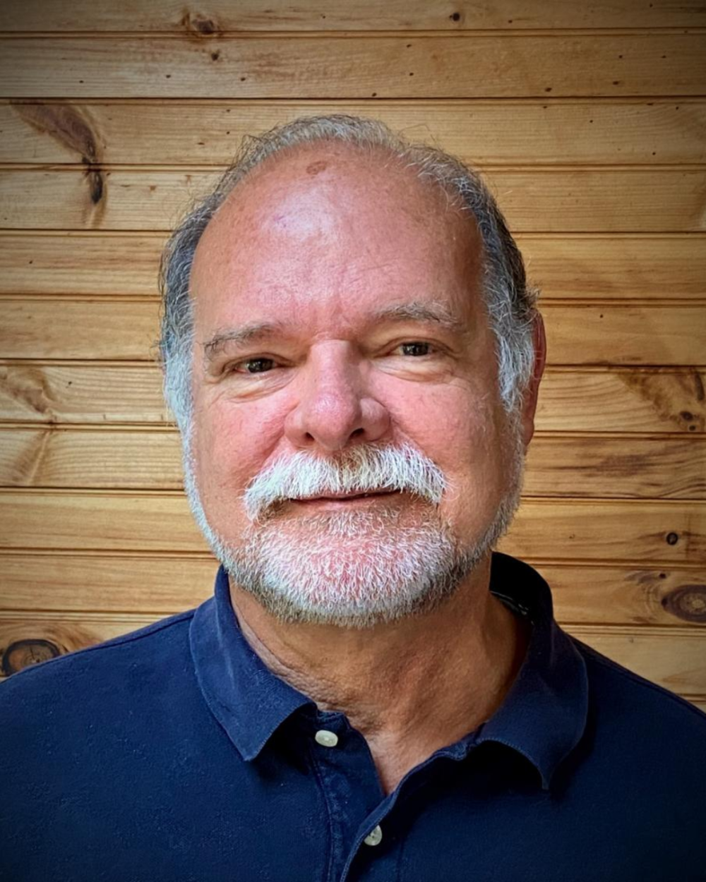 Photo of Dr. Robert Siegel, a man with grey hair and a white beard, wearing a blue collared shirt. He stands in front of a wood paneled wall.