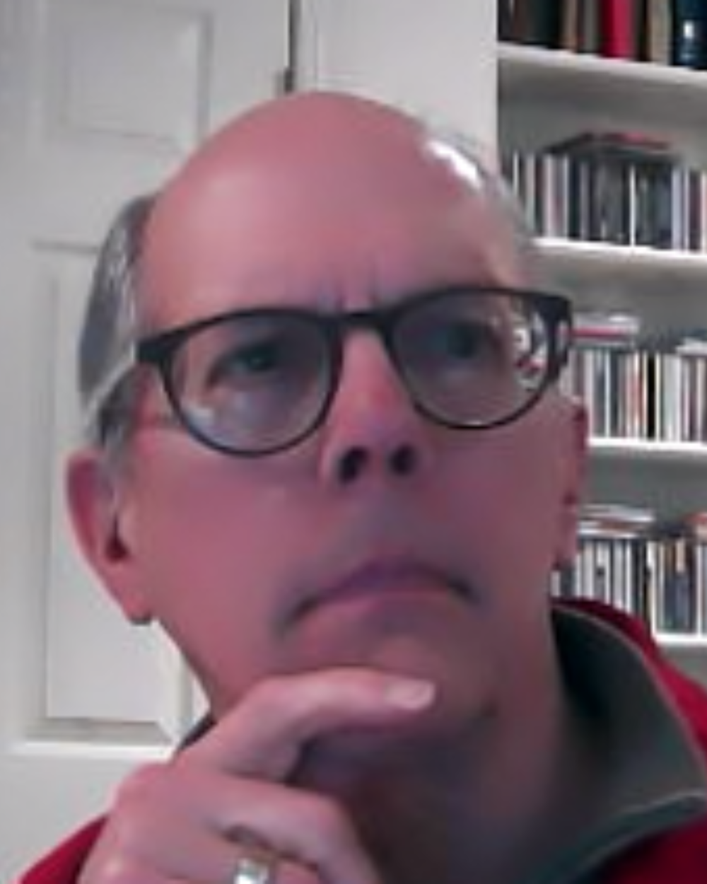 Photo of Dr. Roger Reeves, a man with grey hair, wearing black glasses and a red zip-up sweatshirt. He sits in front of a white door and a bookshelf.