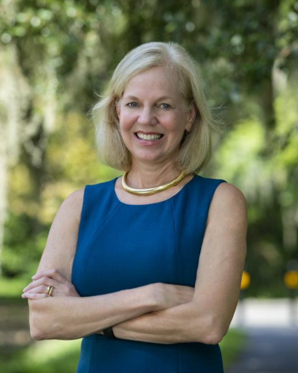 Photo of Dr. Sonja Rasmussen, a woman with light blonde hair, wearing a blue dress. She stands outside with trees in the background.