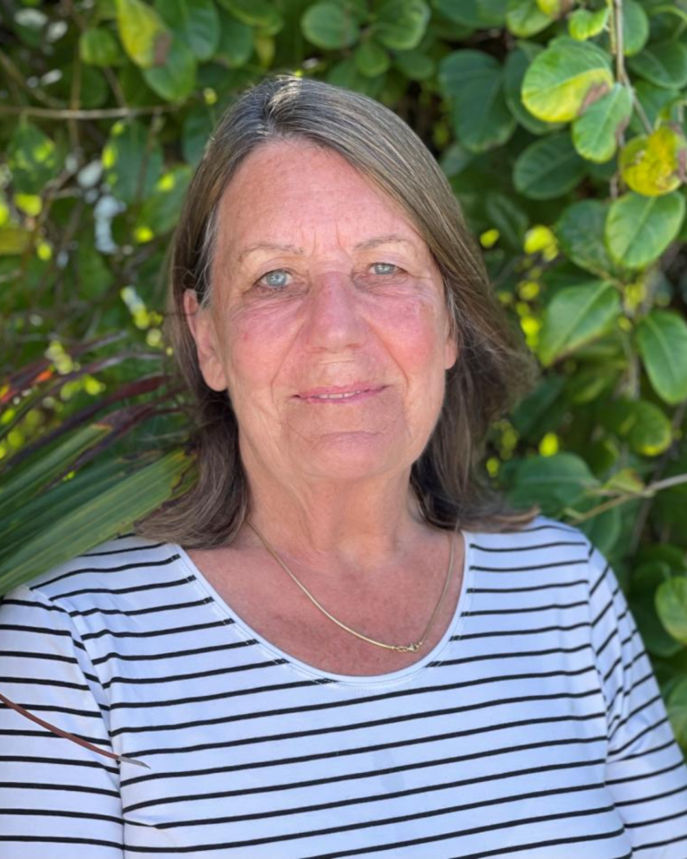 Photo of Betsy Goodwin, a woman with grey-brown hair, wearing a white shirt with stripes. She stands outside in front of a large, green plant.
