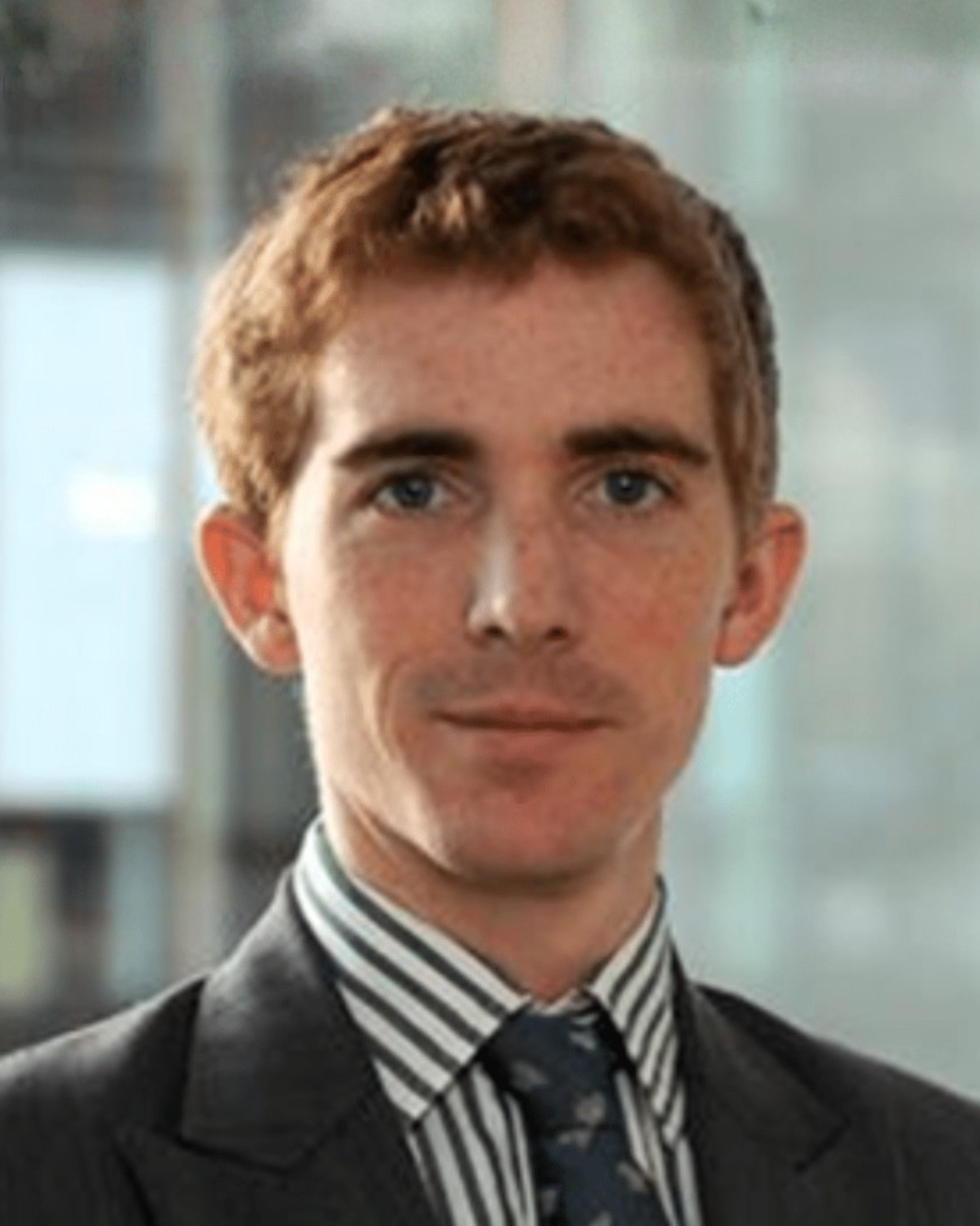 Headshot of Hector Guinness, a man with orange hair, wearing a white collar shirt with grey stripes, a grey suit jacket, and a tie.