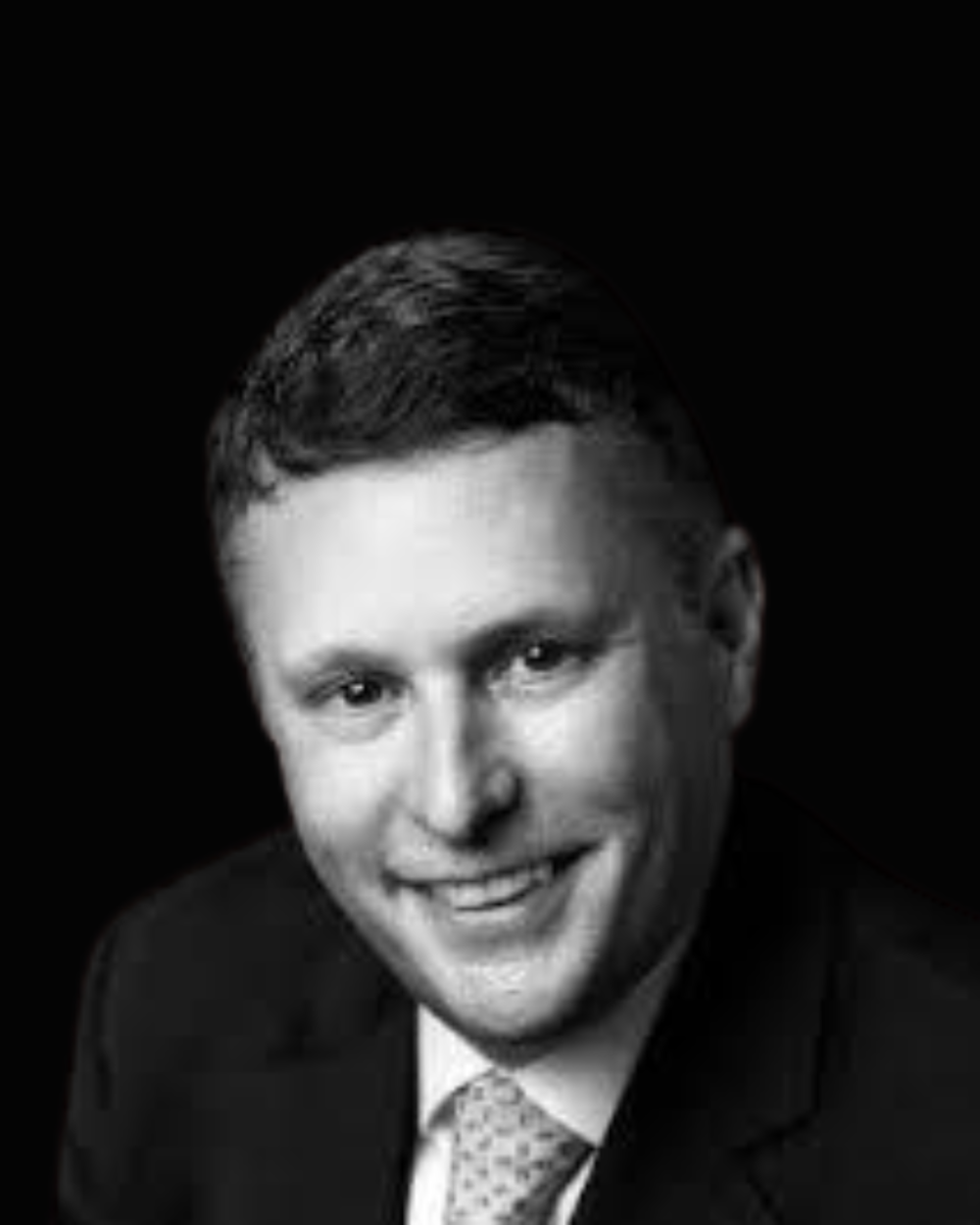 Black and white headshot of John Short, a man wearing a white collar shirt, a tie with small polka dots, and a suit jacket.