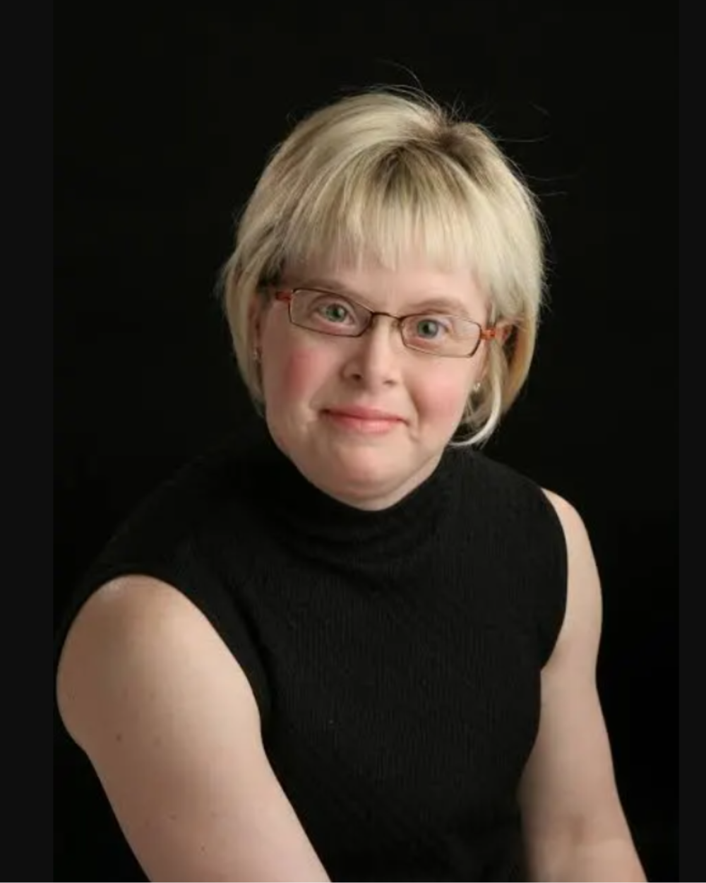 Headshot of Karen Gaffney, a woman with Down syndrome. She has blonde hair and wears pink glasses and a black tank top.
