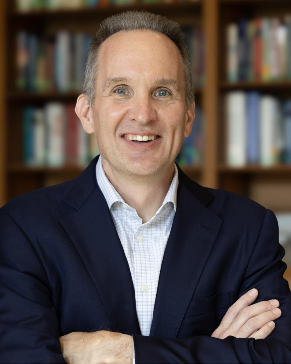 Photo of Michael Kulma, a man with grey hair, wearing a white collar shirt and a black suit jacket. He stands in front of a bookshelf.