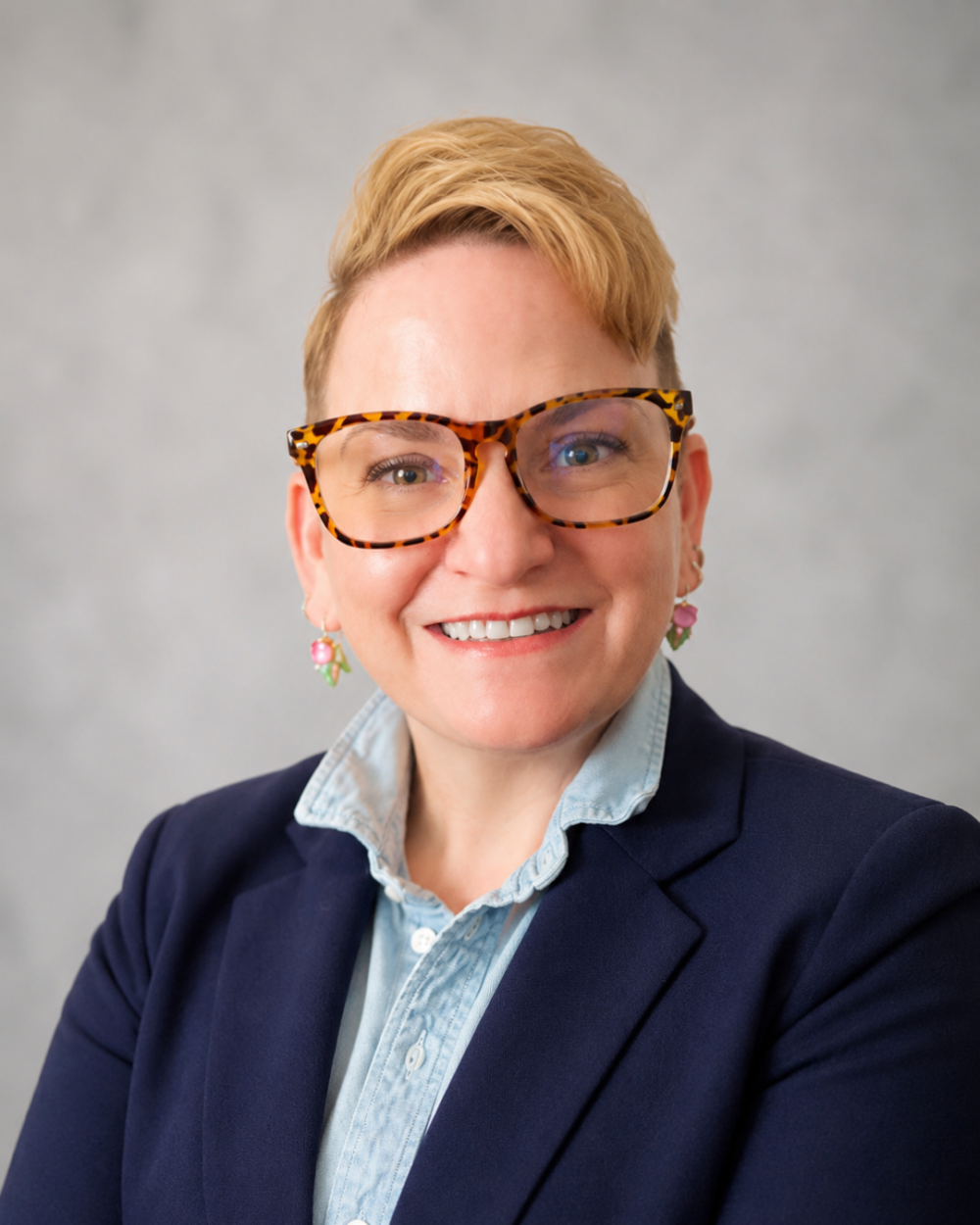 Headshot of Amy Shapley-Farrell, a woman with short blonde hair, multicolored glasses, a blue button-up shirt, navy blazer, and floral earrings.
