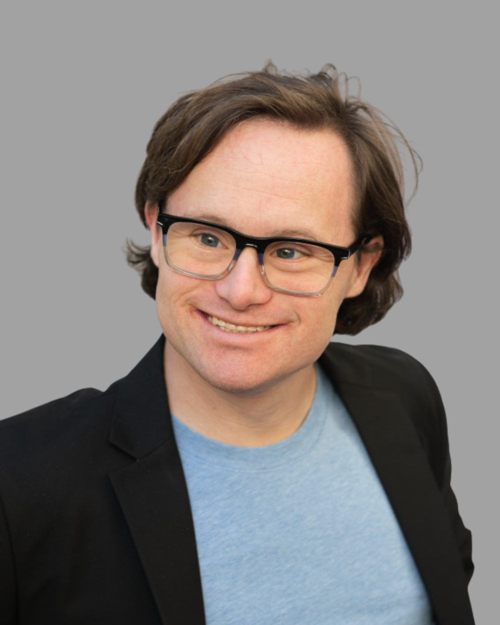 Headshot of Benjamin Golden, a smiling man with Down syndrome with wavy brown hair and dark-framed glasses, wearing a black blazer over a light blue T-shirt. The background is grey.