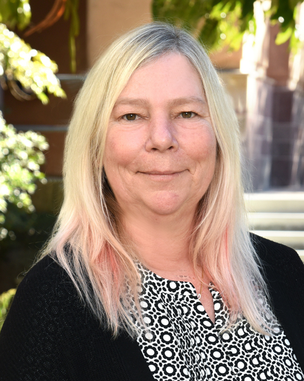 Headshot of Dr. Head, a woman with long blonde and pink hair, smiling gently, wearing a black cardigan over a black-and-white floral patterned top. She is photographed outdoors with greenery and a stone building visible in the background.