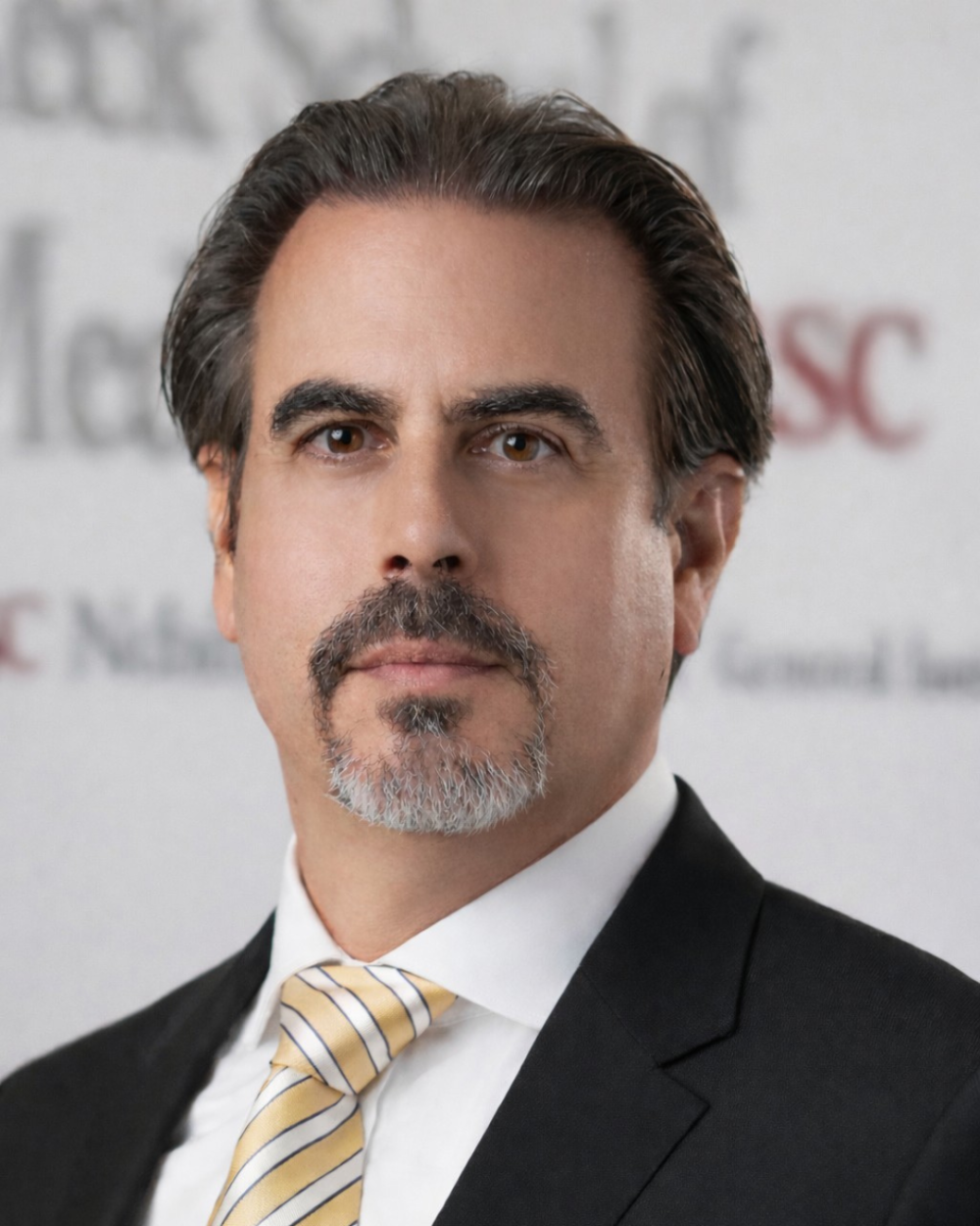 Headshot of Dr. Rafii, a man with dark hair combed back and a gray goatee, wearing a black suit, white dress shirt and a gold-and-white striped tie. He is photographed in front of a blurred backdrop with red text reading "USC" and partial text reading "Keck School of Medicine."