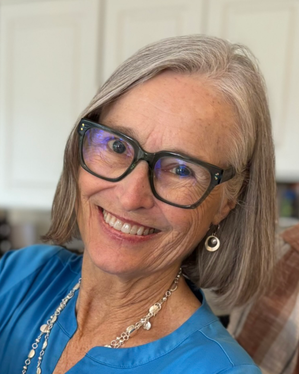 Headshot of Kristi Golden, a smiling woman with shoulder-length gray hair wearing dark rectangular glasses, a blue top, layered silver necklaces and dangling silver earrings. She is photographed indoors in front of white paneled cabinets.