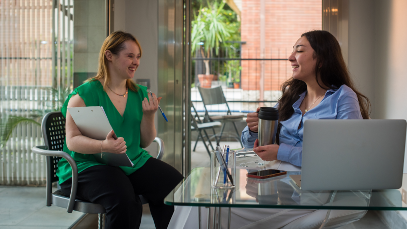 A woman with Down syndrome holding a tablet speaks to another woman at her desk