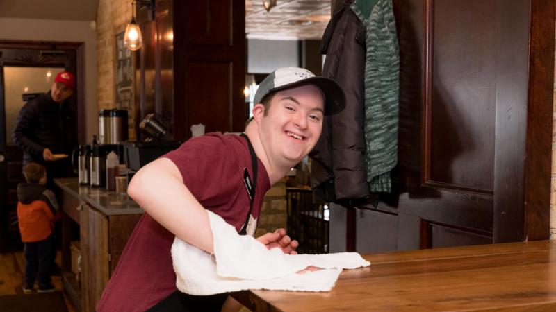 Man with Down syndrome, wearing an apron, smiles while wiping down a table at work