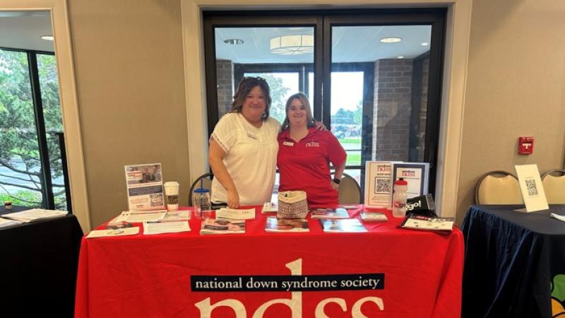A woman with Down syndrome, wearing a red shirt, and a woman wearing a white shirt stand at a table with a red NDSS table cloth.