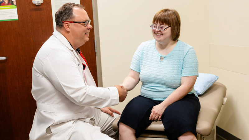 A woman with Down syndrome sits on a seat in a doctor's office. She shakes her doctor's hand.
