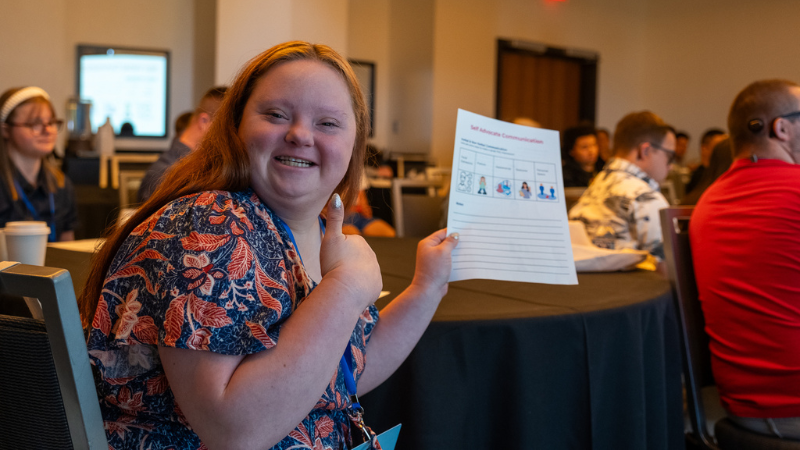 A woman with Down syndrome holds up a piece of paper and gives a thumbs up