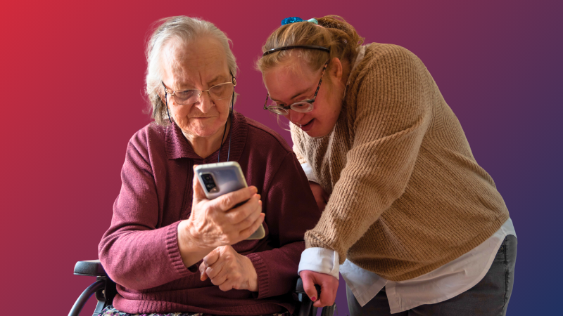 An older woman with Down syndrome looks at a smart phone with an elderly woman sitting in a walker