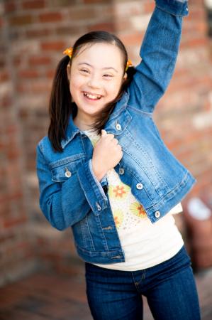 A girl with Down syndrome celebrates with her hand raised. She has pigtails with orange hair ties and wears a denim jacket.