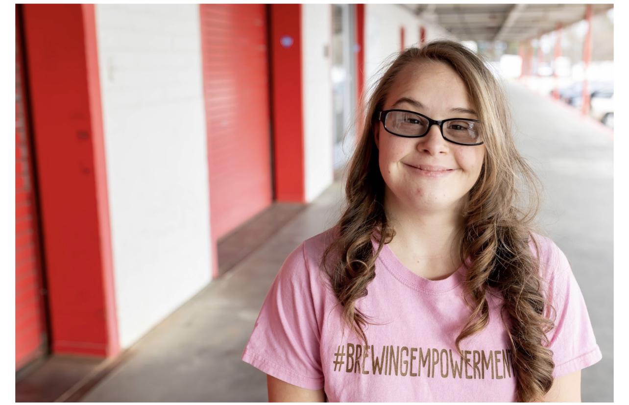 women with down syndrome stands and smiled at the camera. she wears a pink shirt and has long brown curly hair