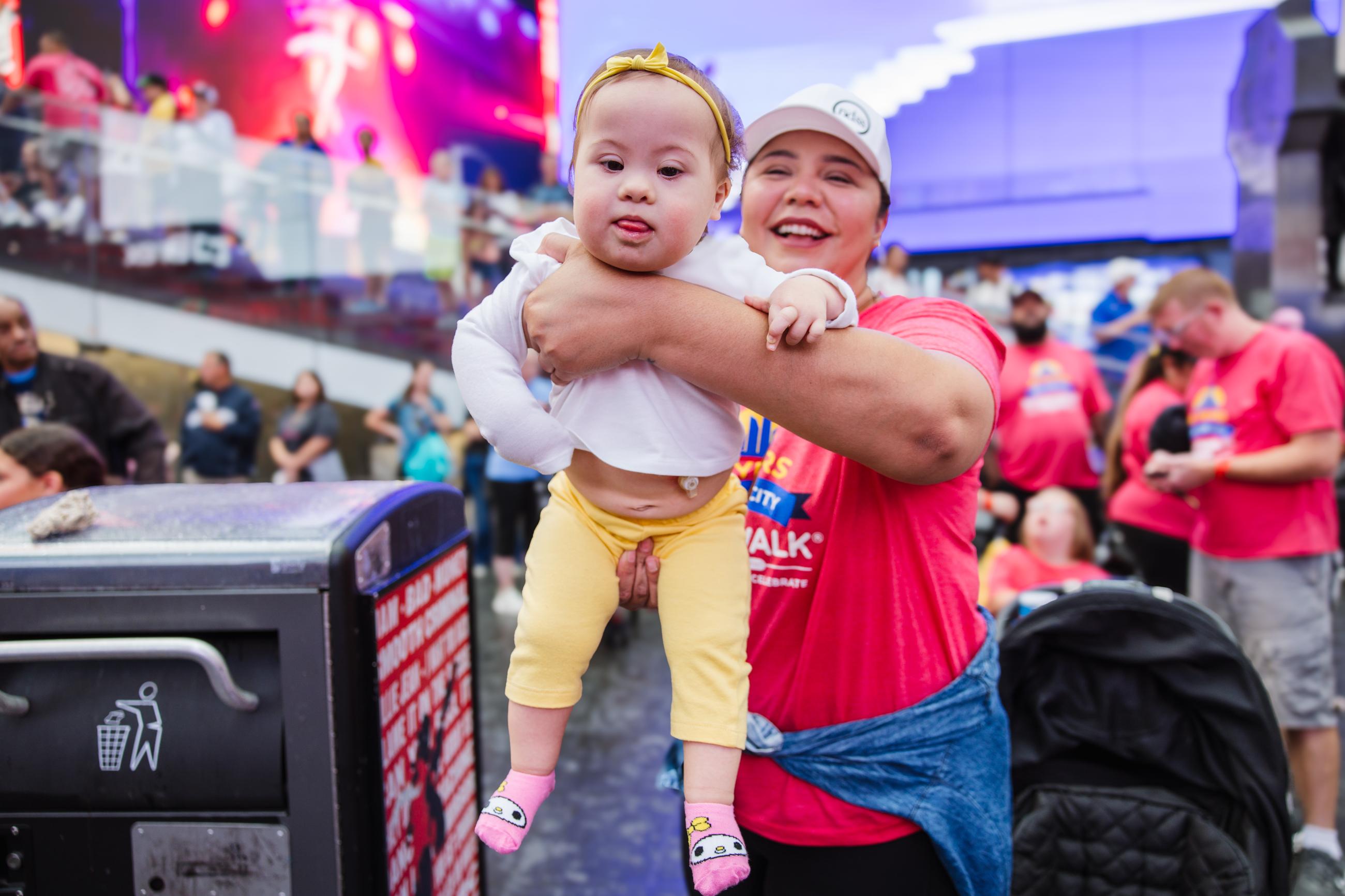 Smiling woman wearing a red Buddy Walk shirt holds up her baby with Down syndrome and a visible G-tube to see the billboard in Times Square