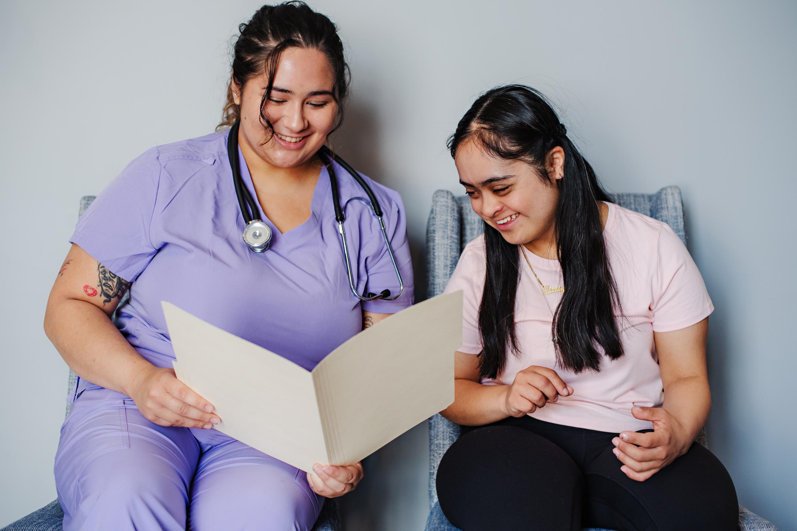 A healthcare professional in lilac scrubs and a woman with Down syndrome review a folder