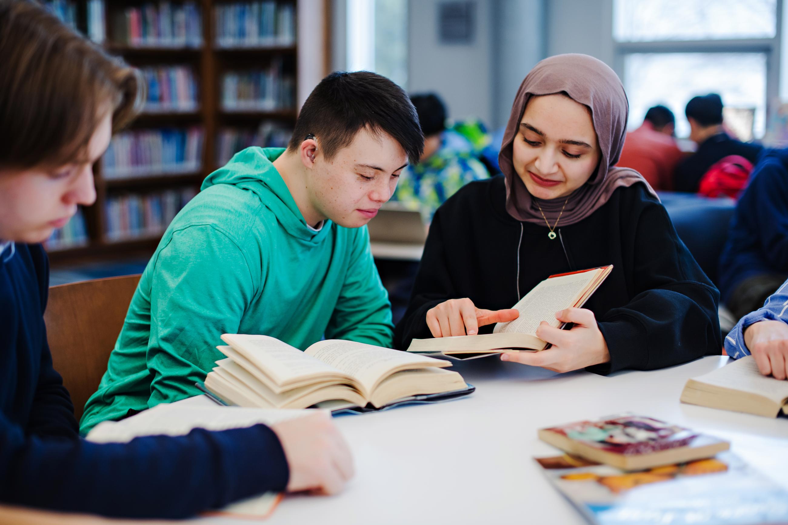 A boy with Down syndrome, wearing a green sweatshirt, reads a book with a girl wearing a hijab