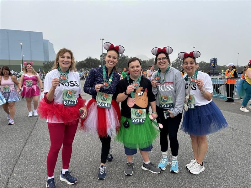 Team NDSS smiles, wearing their NDSS shirts, tutus, and medals. David, a runner with Down syndrome, stands in the middle of the group.