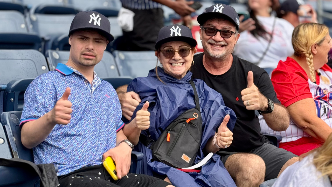 A man with Down syndrome and his family give a thumbs up at Yankee Stadium. They wear New York Yankee hats.